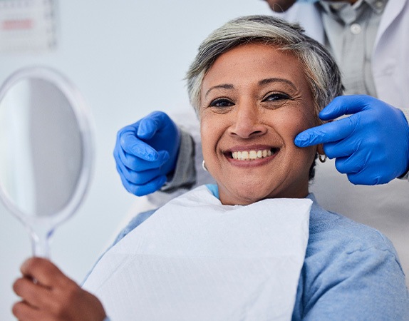 Woman in dental chair smiling holding mirror with dentist in blue gloves touching her cheek